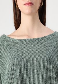 Groene gebreide trui met een losse, off-shoulder halslijn. De textuur is zacht en lichtelijk gestructureerd, met een subtiel patroon. Eenvoudig ontwerp.