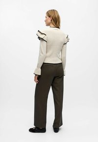 Pull en maille beige clair avec un col et des poignets volants, associé à un pantalon noir ajusté de couleur vert foncé et des chaussures noires sans lacets.