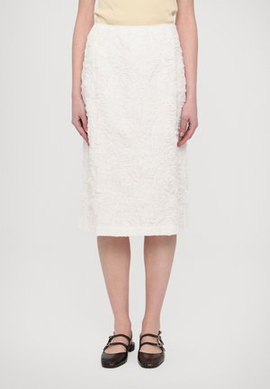 Mujer con una falda blanca texturizada hasta la rodilla con patrón floral y zapatos planos negros de doble tira, de pie contra un fondo blanco.