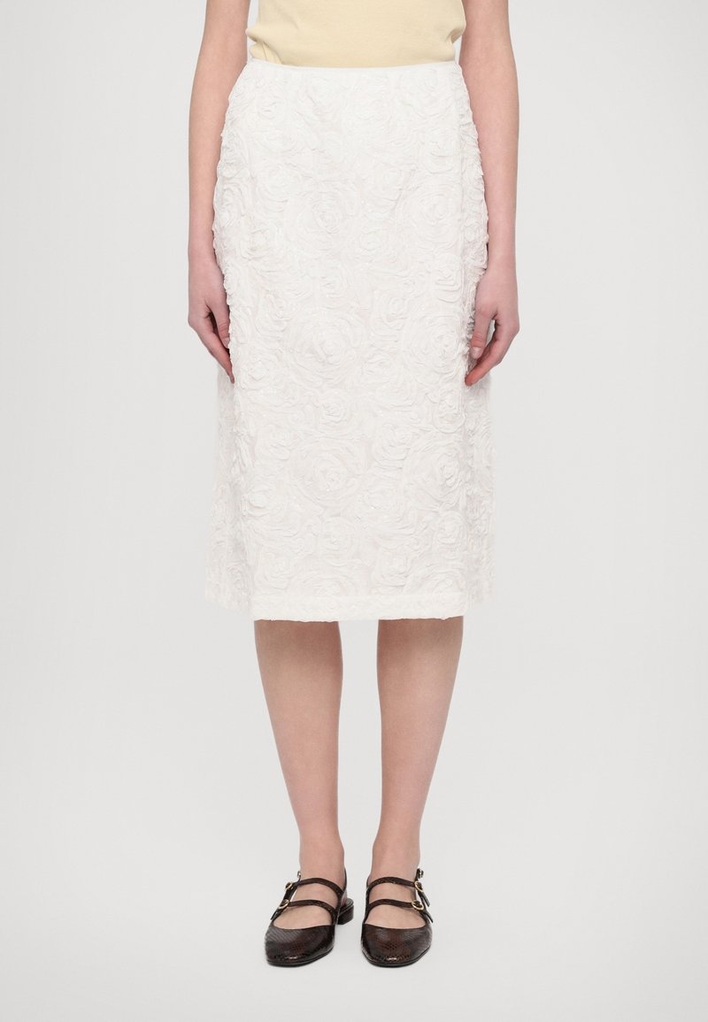 Femme portant une jupe blanche texturée longueur genou avec motif floral et des chaussures plates noires à double bride, debout devant un fond blanc.
