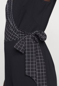 Musta kangasvaate, jossa on kuviollinen solmio kontrastivalkoisessa ruutukuosisessa designissa. Solmio on siististi solmittu vyötärölle.