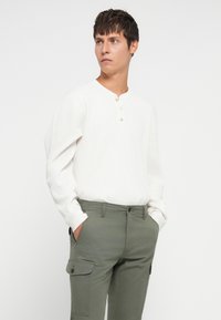 Chemise blanche à manches longues côtelées avec un col henley et deux accents de boutons, associée à un pantalon cargo vert avec des poches latérales.