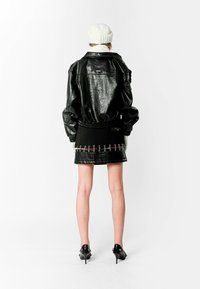 Chaqueta bomber de piel negra con mangas abullonadas, combinada con una falda negra con acentos metálicos. La modelo lleva un gorro de punto blanco.