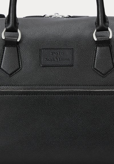 Sac à main en cuir noir texturé avec une poche zippée en argent sous un écusson logo et deux poignées robustes attachées par des anneaux en argent.
