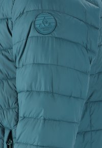 Teal pufferjakke med quiltet design, et rundt Whistler Danmark-logo og lynlås lukning i bunden. Glat, letvægtsstof.