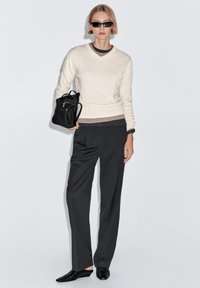 Kvinde iført sorte solbriller, cremefarvet sweater over lagdelte toppe, mørkegrå bukser, sorte sko, holder en sort læderrygsæk mod en hvid baggrund.