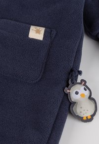 Marineblaues Fleece-Kleidungsstück mit strukturierter Oberfläche, ausgestattet mit einer aufgesetzten Tasche und einem Stoffetikett in Form einer Eule mit kontrastierenden Farben.