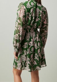 Abito floreale verde con maniche lunghe, caratterizzato da una vita stretta, motivi di fiori rosa e un tessuto leggero e fluente.