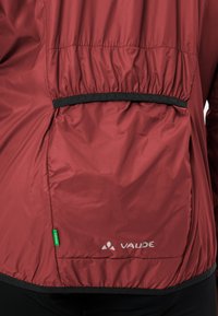 Giacca Vaude rossa sul retro con vita elastica, rifiniture nere e un'etichetta verde "Green Shape" vicino al lato sinistro in basso.
