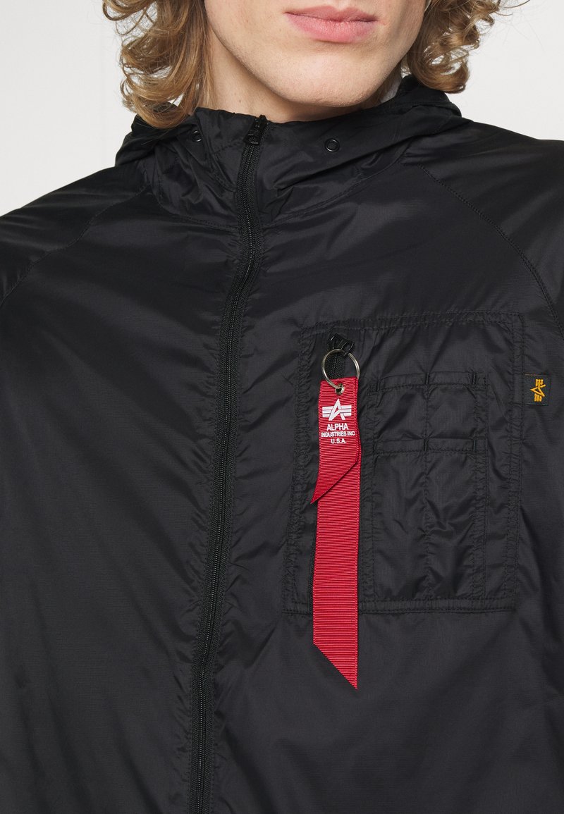 Schwarze leichte Jacke mit hohem Kragen, ausgestattet mit einer Patte-Tasche, einem Metallreißverschluss und einem roten Etikett mit Branding. Glatte Textur.