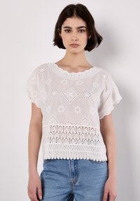 White cotton blouse featuring embroidered floral patterns, a scalloped neckline, and lace detailing at the hem. Worn with blue jeans.