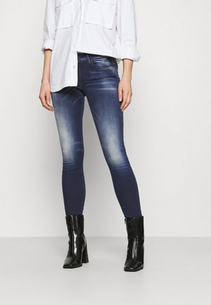 Jeans skinny de color azul oscuro con ligeras desgastes en los muslos, combinados con una camisa blanca de botones y botas de tacón negras.