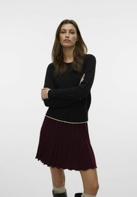 Svart långärmad cardigan med knappar, i kombination med en vinröd plisserad kjol. Gräddfärgad kantning i midjan, texturerad tyg, knä-lång fåll.