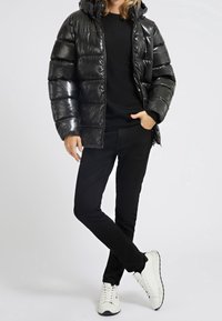 Černá lesklá péřová bunda s kapucí, se šitými sekcemi, v kombinaci s černými kalhotami a bílými teniskami. Neutrální pozadí.