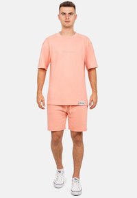 T-shirt rosa chiaro a vestibilità morbida con maniche corte, abbinato a pantaloni corti coordinati. Entrambi presentano ricami minimi e scarpe da ginnastica bianche.