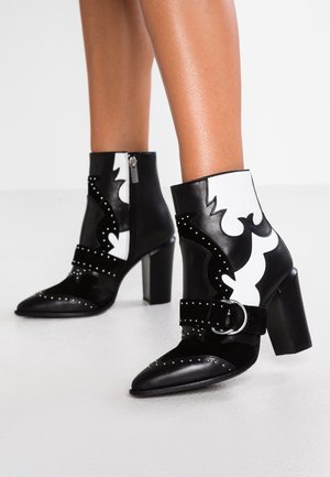 Schwarz-weiße Leder-Stiefeletten mit Blockabsätzen, silbernen Nieten, dekorativen Schnallriemen und seitlichen Reißverschlüssen an den unteren Beinen einer Person.