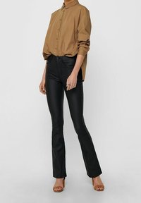 Brązowa koszula z kołnierzykiem, zapinana na guziki, włożona w wysokie czarne spodnie z imitacji skóry o rozszerzanym kroju, noszona z beżowymi sandałami na blokowej podeszwie.