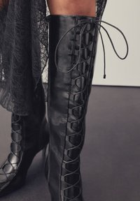 Stivali neri alti fino al ginocchio in pelle lucida con design frontale allacciare e tacco a spillo stretto. Cerniera laterale per facilitare la calzata.