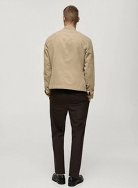 Beige suede jacket with a simple design, worn over brown trousers and black shoes. Features a standard collar and clean lines.