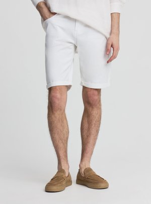 Mężczyzna ubrany w białe szorty z podwiniętym brzegiem, brązowe buty wsuwane i biały longsleeve, stoi z jedną ręką w kieszeni.