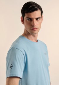 Camiseta de algodón azul claro con cuello redondo y mangas cortas. Presenta un pequeño logo en el pecho y un emblema negro en la manga izquierda.