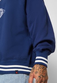 Marineblå sweatshirt med rund halsudskæring, ribbede manchetter og stribede hvide detaljer i bunden. Synlig tatoveret hånd, der holder tøjet.