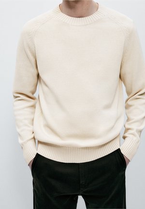 Man wearing a cream-colored knit sweater with ribbed cuffs and hem, hands in pockets, dressed in black pants against a plain background.