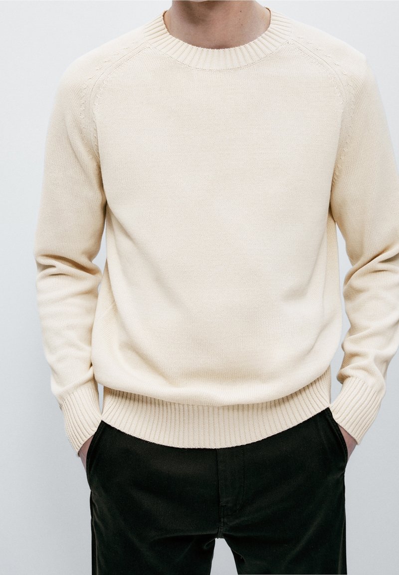 Homme portant un pull tricoté de couleur crème avec poignets et ourlet côtelés, mains dans les poches, habillé d’un pantalon noir sur fond uni.