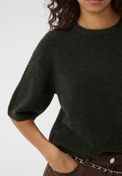 Femme aux cheveux bouclés portant un pull vert foncé pelucheux et un pantalon sombre avec une ceinture chaîne argentée, main dans la poche, sur un fond uni.
