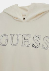 Tejfehér kapucnis felső, elülső dizájnján a "GUESS" felirat ezüst strasszokkal. Puha anyag, laza fazon és állítható kapucni.