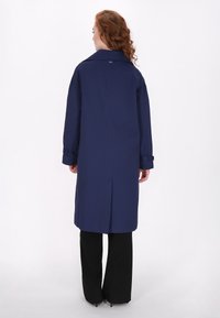 Marineblauer Trenchcoat mit klassischem Kragen, langen Ärmeln und einem Rückenbelüftungsbereich, kombiniert mit schwarzen Hosen. Glatte Textur, minimale Details.