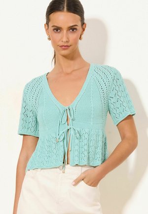 Žena nosi mintovo zelený krátky pletený kardigán so zaväzovaním, skombinovaný s bielymi nohavicami s vysokým pásom, stojaca na svetlom pozadí.