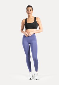 Brassière de sport noire, leggings violets taille haute, texture lisse, design ajusté, associée à des chaussures de sport blanches, posant devant un fond neutre.