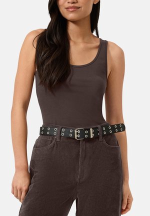 Vrouw draagt een bruin tanktop, hoog getailleerde bruine corduroy broek en een zwarte riem met zilveren oogjes en dubbele gesp.