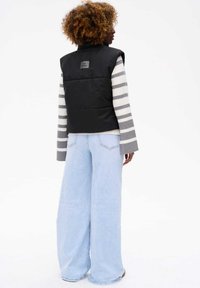 Czarna ocieplana kamizelka z wysokim kołnierzem, noszona na szaro-białym swetrze w paski. Jasnoniebieskie szerokie dżinsy o luźnym kroju.