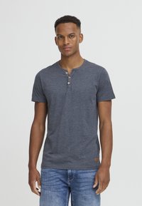 Camiseta henley gris de manga corta, hecha de un tejido suave, con un estampado de tres botones y una pequeña etiqueta de logo marrón en el dobladillo.