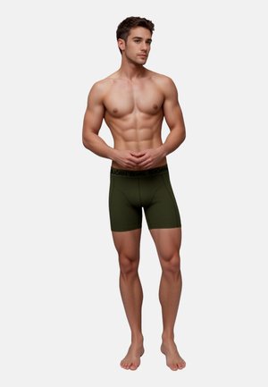 Muskelöser Mann steht barfuß vor weißem Hintergrund, trägt dunkelgrüne eng anliegende Boxershorts mit markiertem Bund, die Hände vor dem Körper verschränkt.