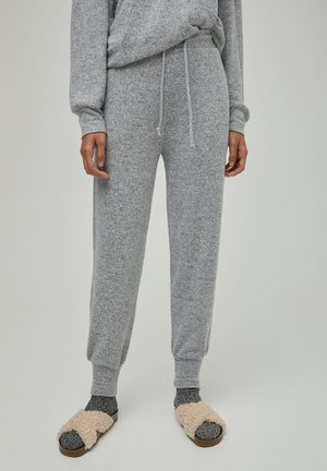 Person iført grå strikkede joggingbukser med en matchende top, mørke sokker og beige, bløde slip-on sandaler, stående mod en ensfarvet baggrund.