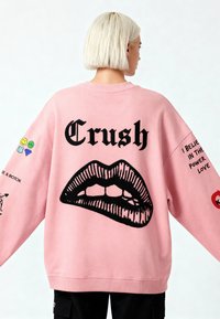 Felpa oversized rosa con grafica nera a forma di labbra, scritta "Crush" e vari patch colorati sulle maniche, esposta su uno sfondo chiaro.