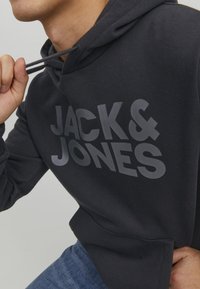 Felpa nera in cotone con un grande e audace logo grigio "JACK & JONES" sul petto, dotata di tasca a marsupio e cappuccio con cordoncino.