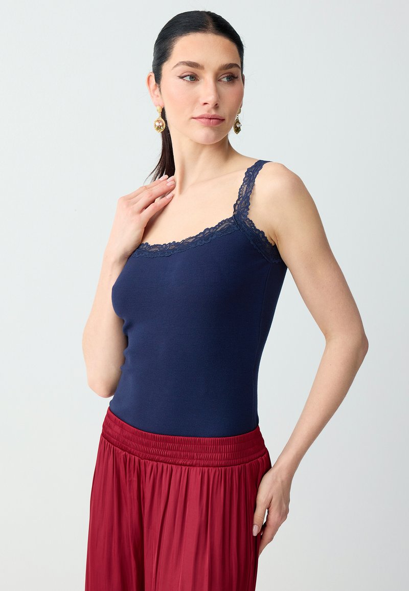 Femme aux cheveux foncés portant des boucles d'oreilles en or, un débardeur bleu marine orné de dentelle et une jupe plissée rouge taille haute, regardant sur le côté.