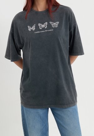 Osoba nosząca wyblakły, czarny, oversize'owy T-shirt z trzema białymi grafikami motyli i napisem „Californian Dreamer” na piersi.