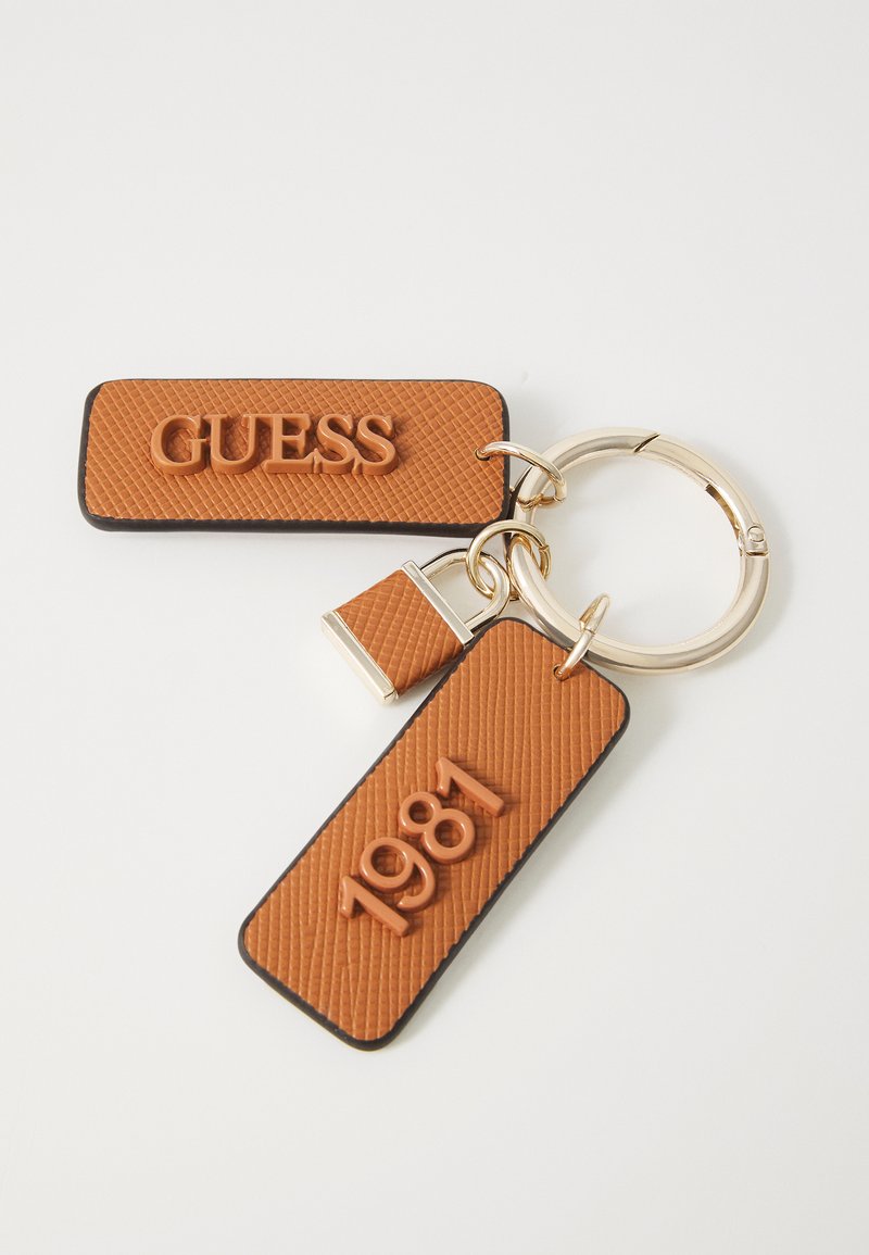 Llaveros marrón con etiquetas de goma texturizada grabadas con "GUESS" y "1981", unidas a un anillo de metal de tono plateado y un detalle de candado.