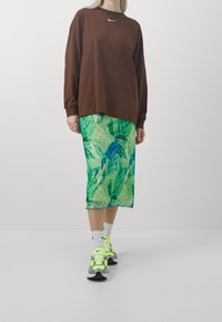 Braunes Langarmhemd mit weißem Logo, grüner Rock mit blauem Batikmuster und graue Sneakers mit neonfarbenen Akzenten und weißen Socken.