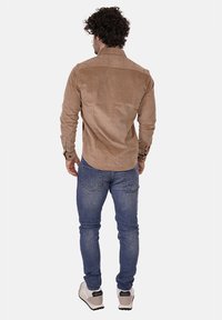 Camicia in velluto a coste marrone con colletto button-down, maniche lunghe e tessuto testurizzato. Abbinata a jeans in denim blu e sneakers di colore chiaro.