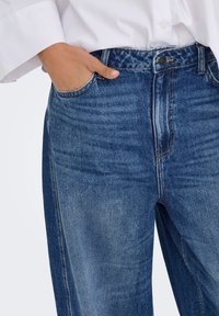 Person wearing blue faded jeans with hand in front pocket and white long-sleeve shirt tucked in at the waist.