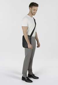Borsa a tracolla nera in materiale sintetico con chiusura a zip, design piatto, logo in rilievo, indossata sulla spalla sinistra da una persona con pantaloni grigi e una camicia bianca.