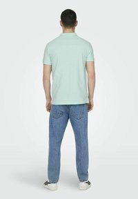 Mintgrünes Polohemd mit kurzen Ärmeln, kombiniert mit hellblauen Jeans aus Denim. Die Jeans haben Gesäßtaschen. Weiße Sneaker vervollständigen den Look.