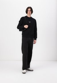 Black sweatshirt with ribbed cuffs, featuring a small logo. Paired with loose black trousers and grey-accents sneakers. Minimalist design.