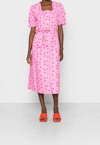 Robe rose avec des manches à volants courtes, décolleté carré et imprimés de fraises. Présente une ceinture à nouer et une fermeture à boutons sur le devant. Sandales à talons rouges.